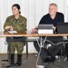 Two individuals are seated at a table in what appears to be a briefing or meeting room. One person is wearing Japanese Ground Self-Defense Force (JGSDF) camouflage uniform, and the other is a civilian in a black shirt. A Dell laptop and an additional monitor are set up on the table, suggesting a presentation or working session.