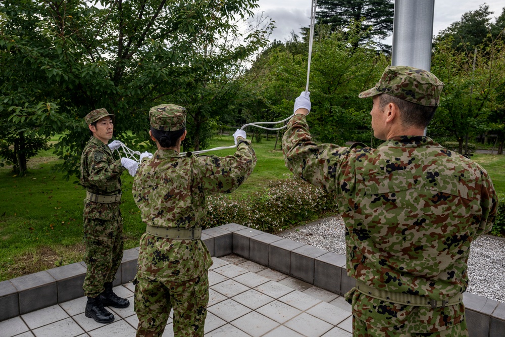 Three military personnel in Japanese Ground Self-Defense Force (JGSDF) camouflage uniforms are conducting a flag ceremony at a flagpole. They are wearing white gloves, suggesting a formal or ceremonial procedure. The setting appears to be a well-maintained outdoor area with trees and a tiled platform.
