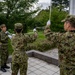 Three military personnel in Japanese Ground Self-Defense Force (JGSDF) camouflage uniforms are conducting a flag ceremony at a flagpole. They are wearing white gloves, suggesting a formal or ceremonial procedure. The setting appears to be a well-maintained outdoor area with trees and a tiled platform.
