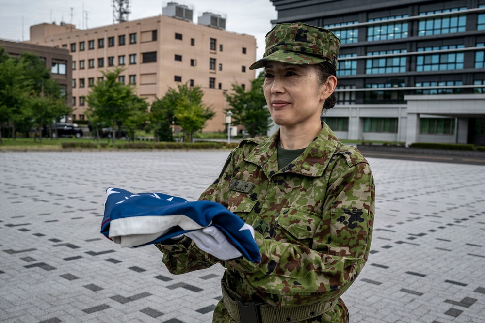 A female military personnel wearing Japanese Ground Self-Defense Force (JGSDF) camouflage uniform is holding a neatly folded flag, identifiable as the United States flag by its visible blue field with white stars and red/white stripes. She is standing in an open paved plaza area with modern buildings and trees visible in the background. The setting appears to be a military or government installation.
