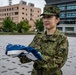 A female military personnel wearing Japanese Ground Self-Defense Force (JGSDF) camouflage uniform is holding a neatly folded flag, identifiable as the United States flag by its visible blue field with white stars and red/white stripes. She is standing in an open paved plaza area with modern buildings and trees visible in the background. The setting appears to be a military or government installation.