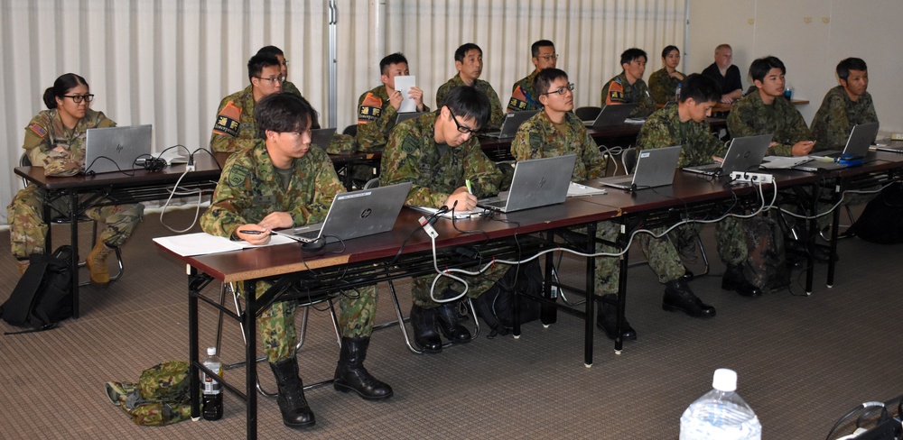 A classroom setting showing military personnel, predominantly in Japanese Ground Self-Defense Force (JGSDF) camouflage uniforms, attending a training or briefing session. Participants are seated at tables equipped with HP laptops and appear to be taking notes. At least one individual in a different uniform, likely a U.S. military member, is visible on the left side.