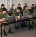 A classroom setting showing military personnel, predominantly in Japanese Ground Self-Defense Force (JGSDF) camouflage uniforms, attending a training or briefing session. Participants are seated at tables equipped with HP laptops and appear to be taking notes. At least one individual in a different uniform, likely a U.S. military member, is visible on the left side.