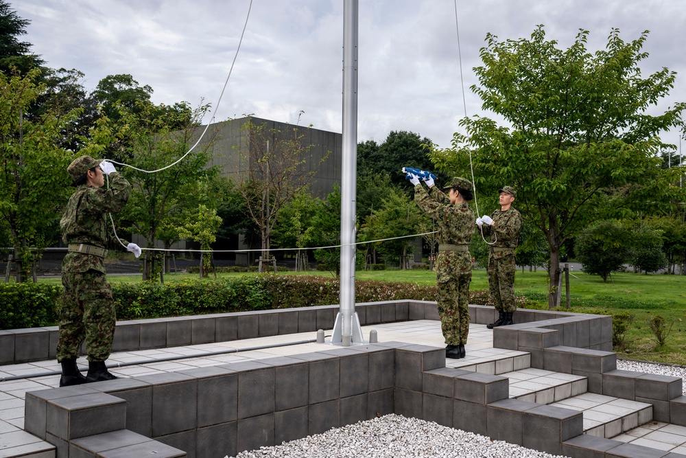 Three military personnel in camouflage uniforms and white gloves are performing a flag ceremony at a flagpole on a tiered stone platform. The soldiers appear to be raising or preparing to raise a folded blue flag, with two handling ropes on either side. The setting appears to be a formal military installation or memorial site surrounded by manicured greenery.