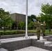Three military personnel in camouflage uniforms and white gloves are performing a flag ceremony at a flagpole on a tiered stone platform. The soldiers appear to be raising or preparing to raise a folded blue flag, with two handling ropes on either side. The setting appears to be a formal military installation or memorial site surrounded by manicured greenery.