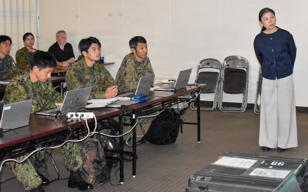 A briefing or training session is taking place in a classroom-style setting, with several individuals in Japanese Ground Self-Defense Force (JGSDF) camouflage uniforms seated at tables with laptops. A civilian woman in a navy blouse and white trousers stands at the front, apparently addressing or presenting to the group. A Western civilian male and a female in civilian attire are also visible seated in the background.