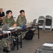 A briefing or training session is taking place in a classroom-style setting, with several individuals in Japanese Ground Self-Defense Force (JGSDF) camouflage uniforms seated at tables with laptops. A civilian woman in a navy blouse and white trousers stands at the front, apparently addressing or presenting to the group. A Western civilian male and a female in civilian attire are also visible seated in the background.