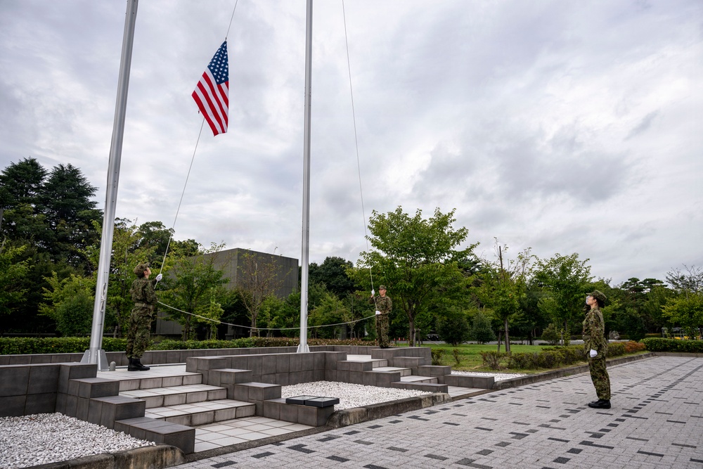 Three military personnel in camouflage uniforms are conducting a flag ceremony at a formal outdoor plaza, raising or lowering a United States flag at half-staff position on a tall flagpole. The setting appears to be a military installation with a structured ceremonial area featuring tiered stone steps, white gravel, and paved walkways. The overcast sky and lush green trees form the backdrop of this formal military observance.