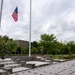 Three military personnel in camouflage uniforms are conducting a flag ceremony at a formal outdoor plaza, raising or lowering a United States flag at half-staff position on a tall flagpole. The setting appears to be a military installation with a structured ceremonial area featuring tiered stone steps, white gravel, and paved walkways. The overcast sky and lush green trees form the backdrop of this formal military observance.