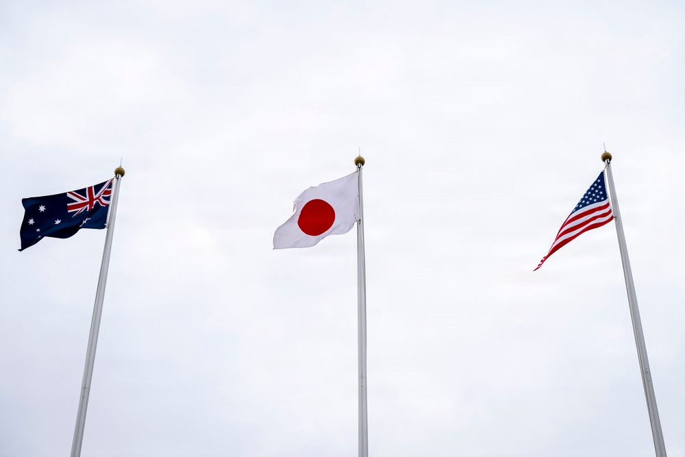 Three national flags are displayed on separate flagpoles against an overcast sky. From left to right, the flags of Australia, Japan, and the United States are visible, all appearing to flutter gently in the wind. The setting suggests a formal diplomatic, governmental, or international event venue.