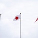 Three national flags are displayed on separate flagpoles against an overcast sky. From left to right, the flags of Australia, Japan, and the United States are visible, all appearing to flutter gently in the wind. The setting suggests a formal diplomatic, governmental, or international event venue.