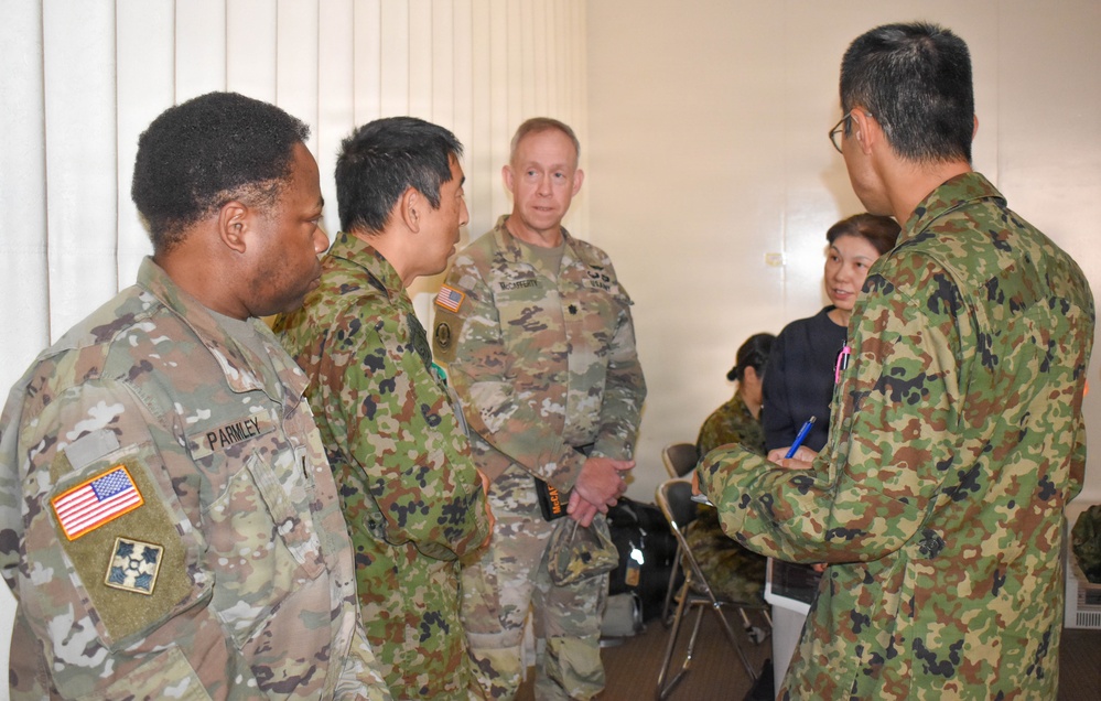 The image shows a group of military personnel from what appear to be two different armed forces engaged in a discussion or briefing in an indoor setting. U.S. Army soldiers wearing OCP (Operational Camouflage Pattern) uniforms are interacting with personnel wearing Japanese Ground Self-Defense Force (JGSDF) camouflage uniforms. The setting suggests a joint military exercise, coordination meeting, or liaison activity.