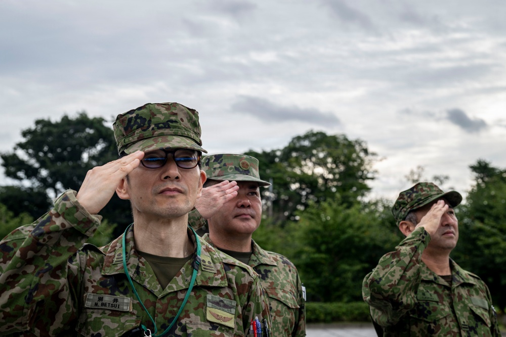 Three military personnel in Japanese Ground Self-Defense Force (JGSDF) camouflage uniforms are performing a formal salute. The foreground soldier has a visible name tag reading 'N. BETSUI' and wears glasses. The scene appears to be an outdoor military ceremony or formation.