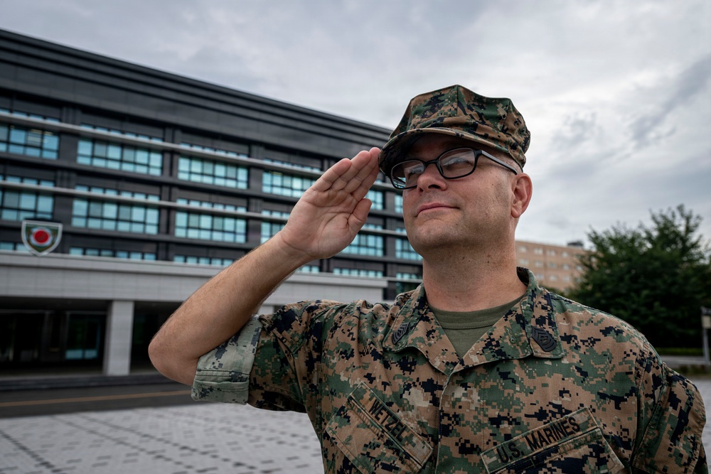 A U.S. Marines service member named Wetzel is photographed rendering a salute in front of a large modern building. The individual is wearing Marine Corps Combat Utility Uniform (MCCUU) in MARPAT woodland camouflage pattern. The setting appears to be a military installation, possibly in Japan based on the visible insignia on the building.