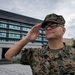 A U.S. Marines service member named Wetzel is photographed rendering a salute in front of a large modern building. The individual is wearing Marine Corps Combat Utility Uniform (MCCUU) in MARPAT woodland camouflage pattern. The setting appears to be a military installation, possibly in Japan based on the visible insignia on the building.