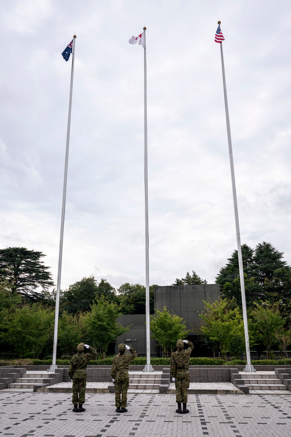 Three military personnel in camouflage uniforms with white gloves stand at salute before three flagpoles displaying the flags of Australia, Japan, and the United States. The ceremony takes place on a paved plaza with tiered stone steps, surrounded by trees and a dark concrete structure in the background. This appears to be a formal tri-national military flag-raising or ceremonial event.