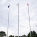 Three military personnel in camouflage uniforms with white gloves stand at salute before three flagpoles displaying the flags of Australia, Japan, and the United States. The ceremony takes place on a paved plaza with tiered stone steps, surrounded by trees and a dark concrete structure in the background. This appears to be a formal tri-national military flag-raising or ceremonial event.