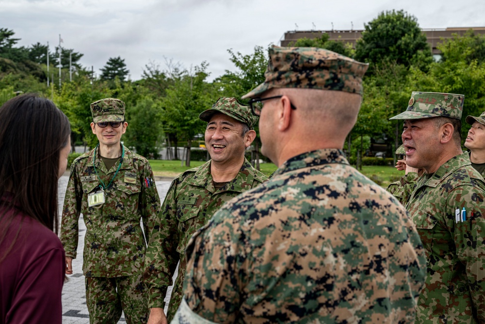 A group of military personnel in two distinct camouflage uniforms are engaged in a friendly outdoor meeting or greeting. The interaction appears to involve personnel wearing Japan Ground Self-Defense Force (JGSDF) uniforms and one individual in what appears to be US Marine Corps MARPAT digital camouflage. The atmosphere is relaxed and cordial, with visible smiling and conversation.