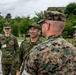 A group of military personnel in two distinct camouflage uniforms are engaged in a friendly outdoor meeting or greeting. The interaction appears to involve personnel wearing Japan Ground Self-Defense Force (JGSDF) uniforms and one individual in what appears to be US Marine Corps MARPAT digital camouflage. The atmosphere is relaxed and cordial, with visible smiling and conversation.