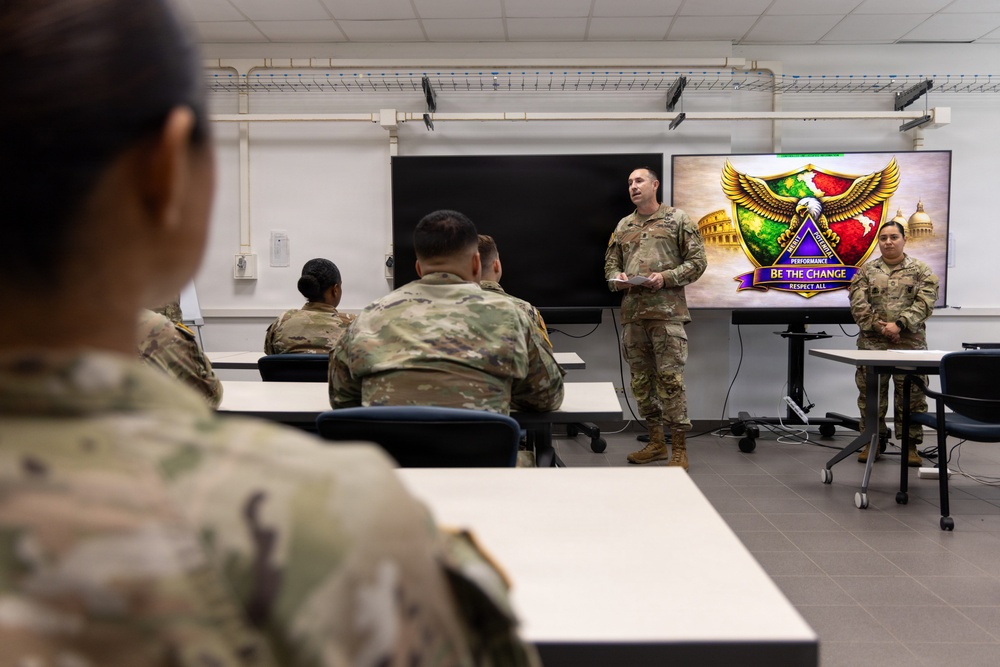 A military briefing or training session is taking place in a classroom setting, with one soldier standing at the front presenting to a group of seated soldiers in camouflage uniforms. A display screen behind the presenter shows a colorful emblem or unit crest with the text 'BE THE CHANGE' and 'RESPECT ALL.' A second soldier stands near the screen, possibly assisting with the presentation.