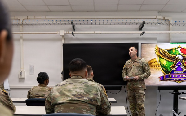 A military briefing or training session is taking place in a classroom setting, with one soldier standing at the front presenting to a group of seated soldiers in camouflage uniforms. A display screen behind the presenter shows a colorful emblem or unit crest with the text 'BE THE CHANGE' and 'RESPECT ALL.' A second soldier stands near the screen, possibly assisting with the presentation.