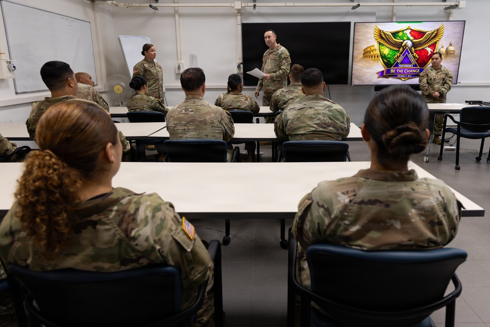 A group of U.S. Army soldiers in OCP (Operational Camouflage Pattern) uniforms are gathered in a classroom setting for a briefing or training session. A male soldier stands at the front holding papers and addressing the group, while a female soldier stands nearby. A display screen on the right shows a unit insignia with the text 'Be The Change' and 'Respect All.'