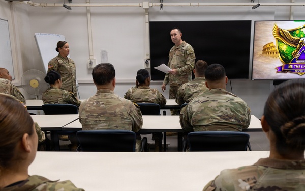 A group of U.S. Army soldiers in OCP (Operational Camouflage Pattern) uniforms are gathered in a classroom setting for a briefing or training session. A male soldier stands at the front holding papers and addressing the group, while a female soldier stands nearby. A display screen on the right shows a unit insignia with the text 'Be The Change' and 'Respect All.'