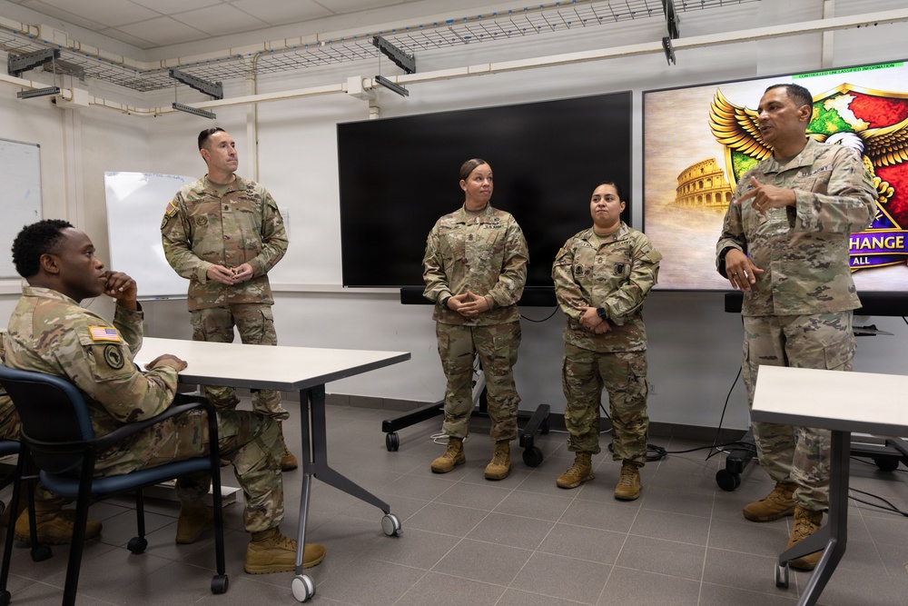 Five U.S. Army soldiers in OCP (Operational Camouflage Pattern) uniforms are gathered in a military briefing or classroom setting. One soldier on the right appears to be speaking or presenting, while the others listen attentively. A large display screen showing a unit insignia with an eagle and the Roman Colosseum is visible in the background, suggesting a unit stationed in or associated with Italy.