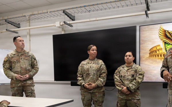 Five U.S. Army soldiers in OCP (Operational Camouflage Pattern) uniforms are gathered in a military briefing or classroom setting. One soldier on the right appears to be speaking or presenting, while the others listen attentively. A large display screen showing a unit insignia with an eagle and the Roman Colosseum is visible in the background, suggesting a unit stationed in or associated with Italy.