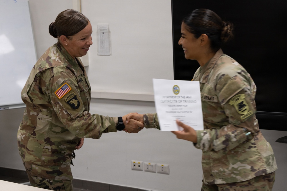 Two female U.S. Army soldiers in OCP (Operational Camouflage Pattern) uniforms are shaking hands during a certificate presentation ceremony. The soldier on the left, wearing a 101st Airborne Division patch and U.S. flag patch, is presenting a Department of the Army Certificate of Training to the soldier on the right. Both individuals are smiling, indicating a positive and celebratory occasion.