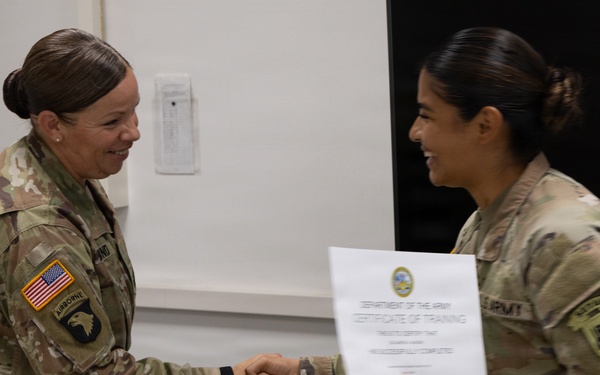 Two female U.S. Army soldiers in OCP (Operational Camouflage Pattern) uniforms are shaking hands during a certificate presentation ceremony. The soldier on the left, wearing a 101st Airborne Division patch and U.S. flag patch, is presenting a Department of the Army Certificate of Training to the soldier on the right. Both individuals are smiling, indicating a positive and celebratory occasion.
