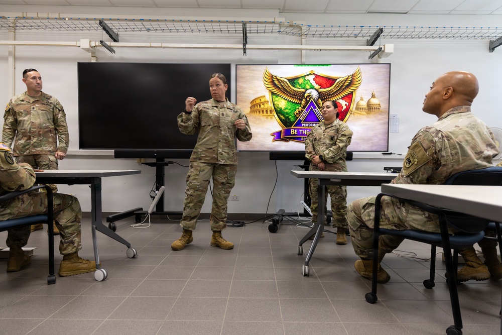 A military briefing or training session is taking place in a modern classroom setting. A female soldier in U.S. Army combat uniform (OCP) is standing and addressing a small group of fellow soldiers. A presentation screen behind her displays what appears to be a unit insignia or patch with an eagle, shield, and the text 'BE THE...' along with imagery of the Colosseum and U.S. Capitol.