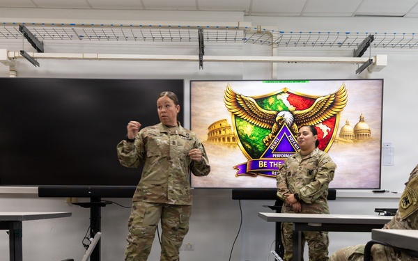A military briefing or training session is taking place in a modern classroom setting. A female soldier in U.S. Army combat uniform (OCP) is standing and addressing a small group of fellow soldiers. A presentation screen behind her displays what appears to be a unit insignia or patch with an eagle, shield, and the text 'BE THE...' along with imagery of the Colosseum and U.S. Capitol.