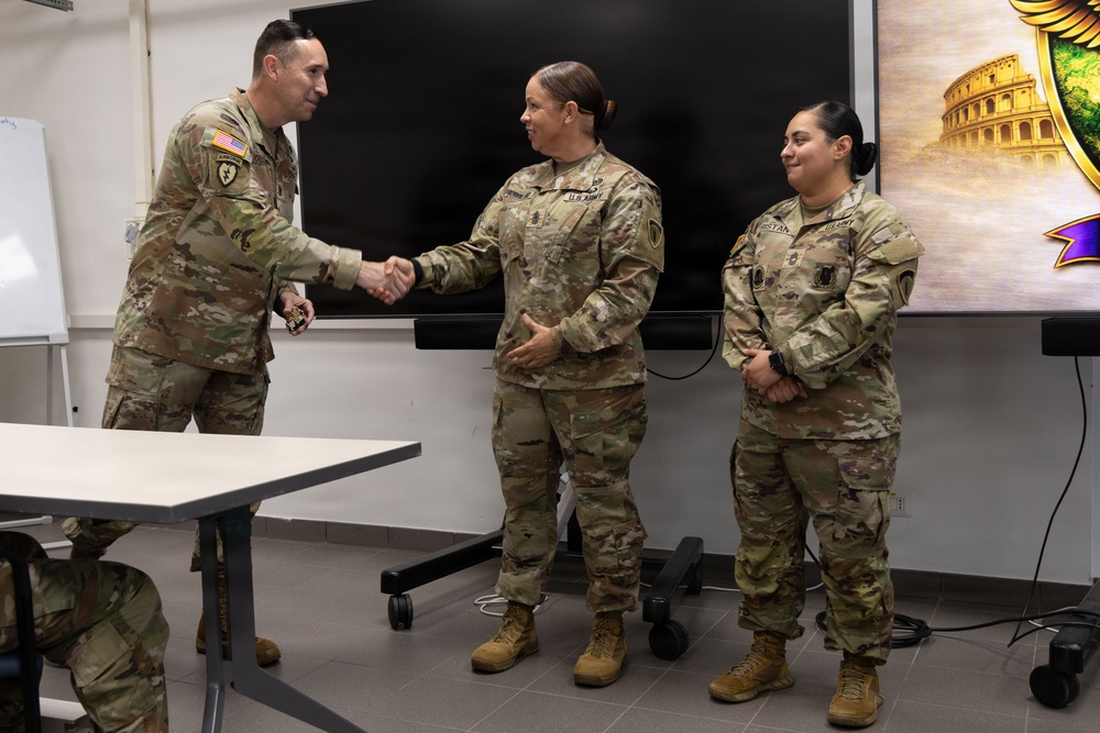 Three U.S. Army soldiers in OCP (Operational Camouflage Pattern) uniforms are gathered in what appears to be a briefing or conference room. A male soldier on the left is shaking hands with a female soldier in the center, suggesting a recognition or award ceremony. A second female soldier stands to the right observing the exchange.