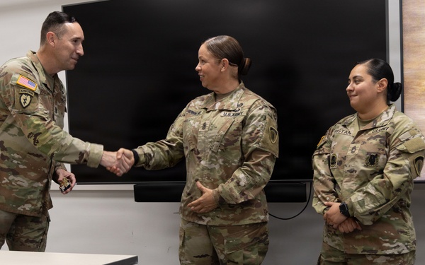 Three U.S. Army soldiers in OCP (Operational Camouflage Pattern) uniforms are gathered in what appears to be a briefing or conference room. A male soldier on the left is shaking hands with a female soldier in the center, suggesting a recognition or award ceremony. A second female soldier stands to the right observing the exchange.