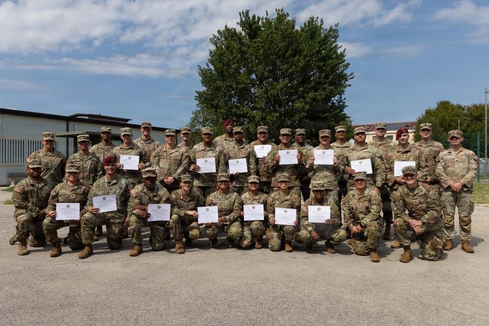 A large group of military personnel in camouflage uniforms are posed together for a group photograph, with most holding certificates or documents. The group is arranged in two rows — a standing back row and a kneeling front row — in an outdoor paved area with buildings and a large tree visible in the background. This appears to be a graduation, award, or recognition ceremony.