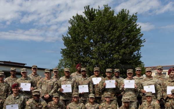 A large group of military personnel in camouflage uniforms are posed together for a group photograph, with most holding certificates or documents. The group is arranged in two rows — a standing back row and a kneeling front row — in an outdoor paved area with buildings and a large tree visible in the background. This appears to be a graduation, award, or recognition ceremony.