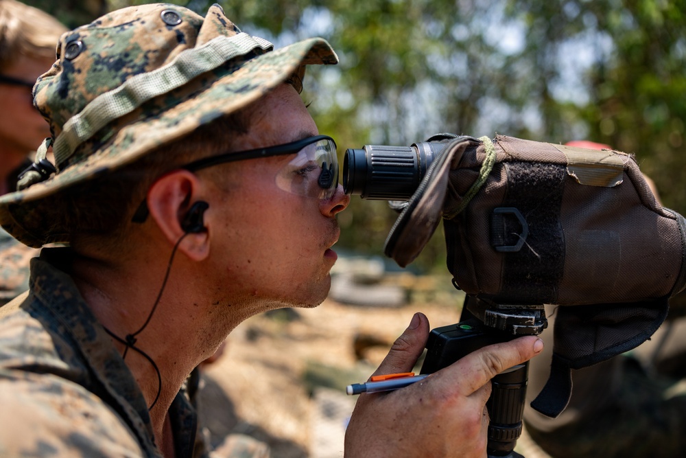 A military personnel member, wearing MARPAT woodland camouflage and a boonie hat, is looking through a spotting scope or monocular optical device mounted on a tripod. The individual is holding a pen and what appears to be a notepad, suggesting they are recording observations. The setting appears to be an outdoor field training or range environment.