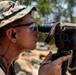 A military personnel member, wearing MARPAT woodland camouflage and a boonie hat, is looking through a spotting scope or monocular optical device mounted on a tripod. The individual is holding a pen and what appears to be a notepad, suggesting they are recording observations. The setting appears to be an outdoor field training or range environment.