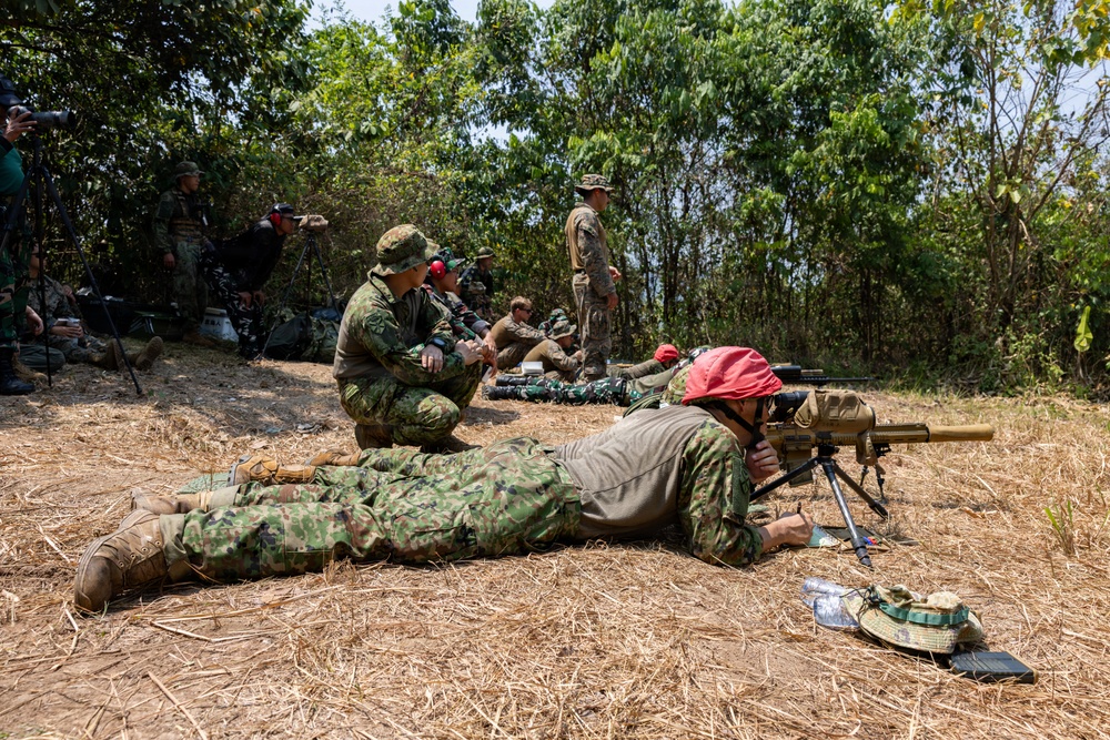 A military training exercise is taking place in an outdoor, wooded area with dry grass terrain. Multiple personnel in camouflage uniforms are engaged in a sniper or marksmanship training session, with one shooter in the prone position aiming a scoped rifle mounted on a bipod. Observers, instructors, and what appear to be media personnel with cameras are present in the background.