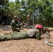 A military training exercise is taking place in an outdoor, wooded area with dry grass terrain. Multiple personnel in camouflage uniforms are engaged in a sniper or marksmanship training session, with one shooter in the prone position aiming a scoped rifle mounted on a bipod. Observers, instructors, and what appear to be media personnel with cameras are present in the background.