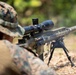 A military personnel in MARPAT (Marine pattern) camouflage uniform is in a prone shooting position with a scoped rifle supported by a bipod. The shooter is aiming downrange in an outdoor environment with natural ground cover. The image appears to depict a marksmanship training or field exercise scenario.