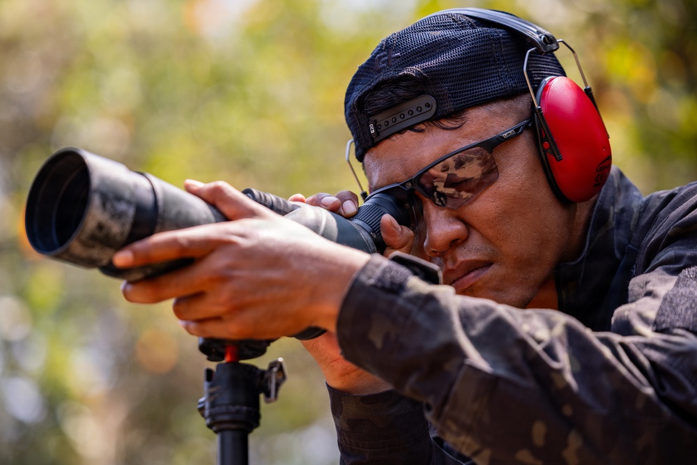 A man in camouflage clothing is looking through a spotting scope or monocular mounted on a tripod in an outdoor setting. He is wearing red ear protection (earmuffs), safety glasses, and a dark baseball cap. The setting and equipment suggest a shooting range or tactical observation activity.