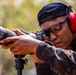A man in camouflage clothing is looking through a spotting scope or monocular mounted on a tripod in an outdoor setting. He is wearing red ear protection (earmuffs), safety glasses, and a dark baseball cap. The setting and equipment suggest a shooting range or tactical observation activity.