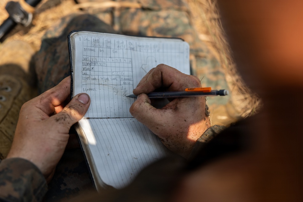A person wearing military camouflage uniform is writing in a small field notebook with a pencil. The notebook contains a structured data table with rows labeled for distance, elevation, wind, calls, and remarks, consistent with a military sniper or shooting data book. The hands are visibly dirty/muddy, suggesting field conditions.
