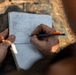 A person wearing military camouflage uniform is writing in a small field notebook with a pencil. The notebook contains a structured data table with rows labeled for distance, elevation, wind, calls, and remarks, consistent with a military sniper or shooting data book. The hands are visibly dirty/muddy, suggesting field conditions.