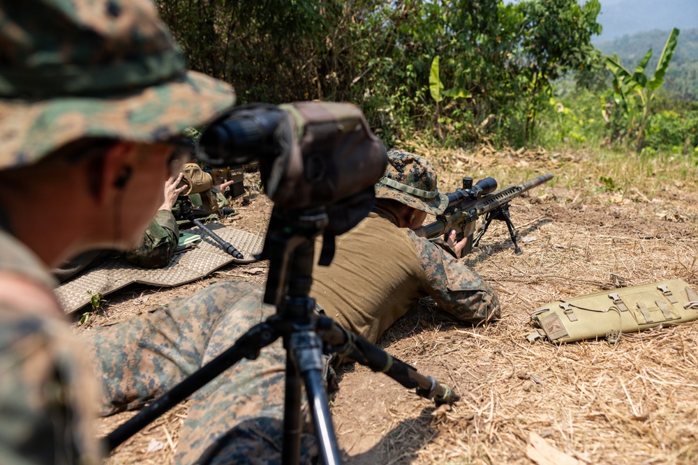 Military personnel in MARPAT (Marine pattern) camouflage uniforms are conducting a sniper or designated marksman training exercise in an outdoor field environment. One shooter is in the prone position behind a scoped rifle mounted on a bipod, while another personnel observes through a spotting scope on a tripod. A third individual is visible in the background, likely recording data or providing support.