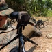 Military personnel in MARPAT (Marine pattern) camouflage uniforms are conducting a sniper or designated marksman training exercise in an outdoor field environment. One shooter is in the prone position behind a scoped rifle mounted on a bipod, while another personnel observes through a spotting scope on a tripod. A third individual is visible in the background, likely recording data or providing support.
