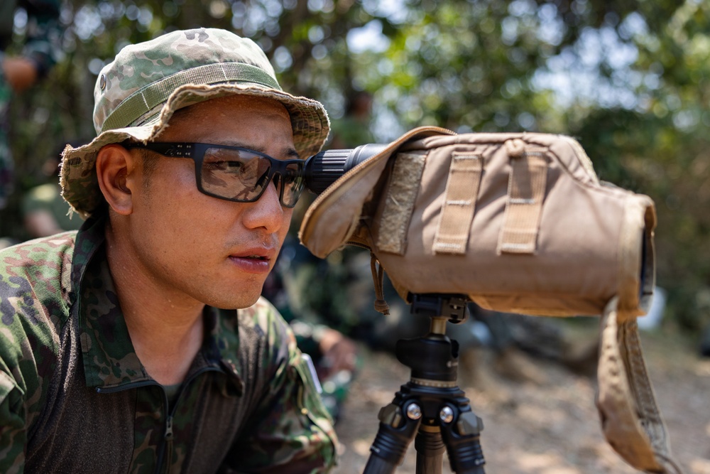 A military personnel wearing camouflage uniform and boonie hat is looking through a spotting scope mounted on a tripod. The scope is covered with a tan/coyote brown protective cover secured with hook-and-loop fasteners. The setting appears to be an outdoor field environment with other personnel visible in the background.