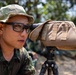 A military personnel wearing camouflage uniform and boonie hat is looking through a spotting scope mounted on a tripod. The scope is covered with a tan/coyote brown protective cover secured with hook-and-loop fasteners. The setting appears to be an outdoor field environment with other personnel visible in the background.